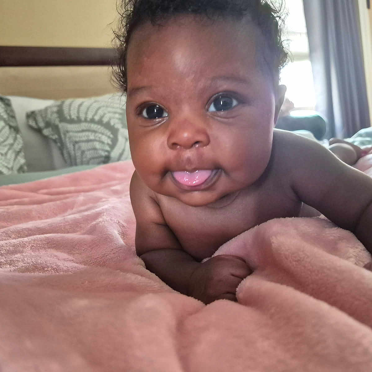 Royal is registered to the contest to win money with this photo: baby, infant, tongue_out, blanket, bed, pillow, indoor, cute, child, face, skin, expression, curly_hair, soft_texture, cozy, room, window, natural_light, portrait, lying_down