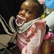 Royal is registered to the contest to win money with this photo: baby, child, pacifier, pink_clothing, headband, flower_accessory, infant, person, holding, cute, indoor, closeup, portrait, hands, soft_toys, blanket, cozy, warm, black_skin, curly_hair