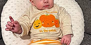 Lylah is registered to the contest to win money with this photo: baby, child, clothing, couch, cute, expression, gray_fabric, hand, indoors, infant, knit, orange, person, pillow, portrait, pumpkin, sitting, smiling_pumpkin, striped_pants, sweater