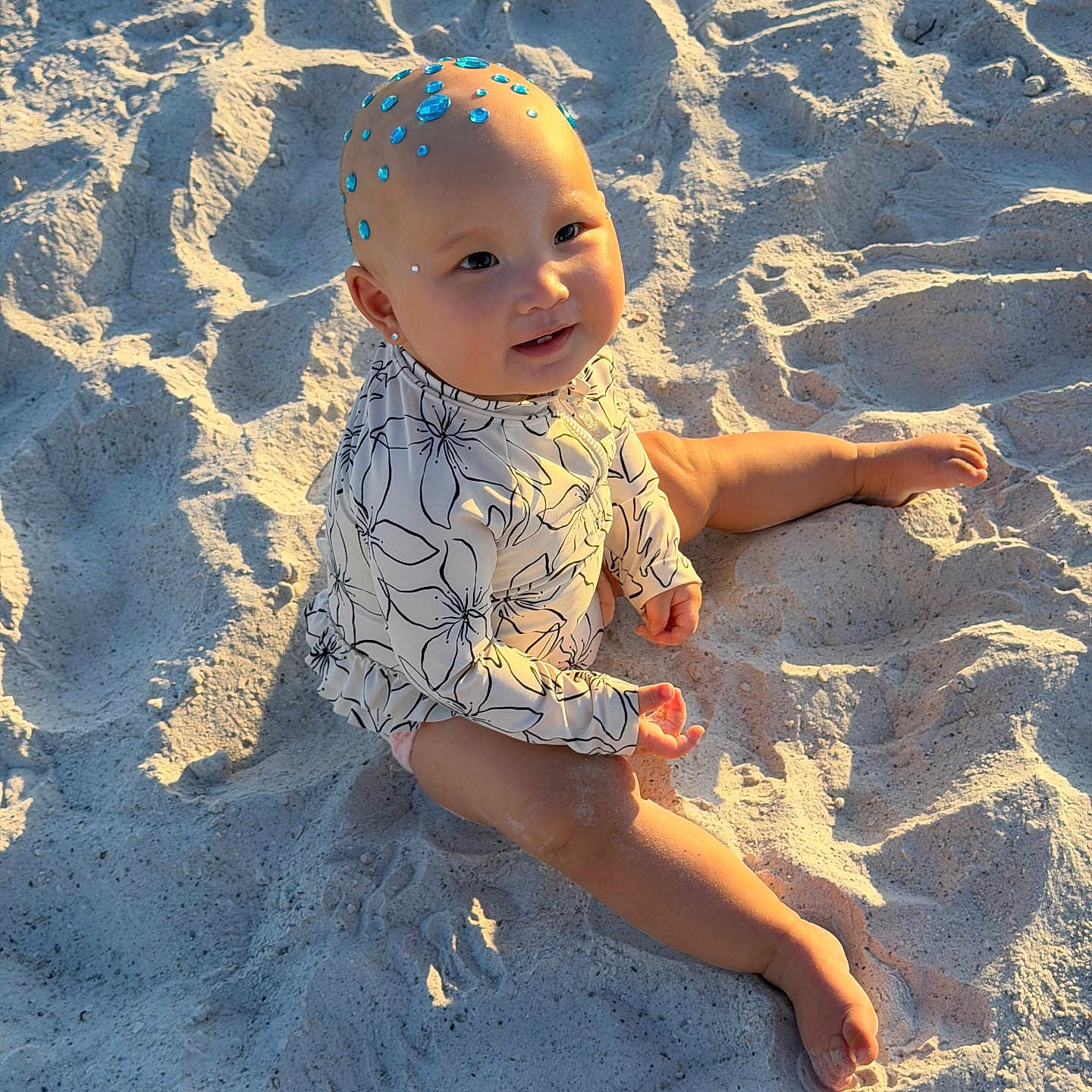 Alana joined the competition — help win amazing prizes! baby, child, sand, beach, sunlight, smile, face, head, gems, skin, barefoot, long_sleeve, floral_pattern, sitting, outdoor, nature, cute, playful, vacation, summer
