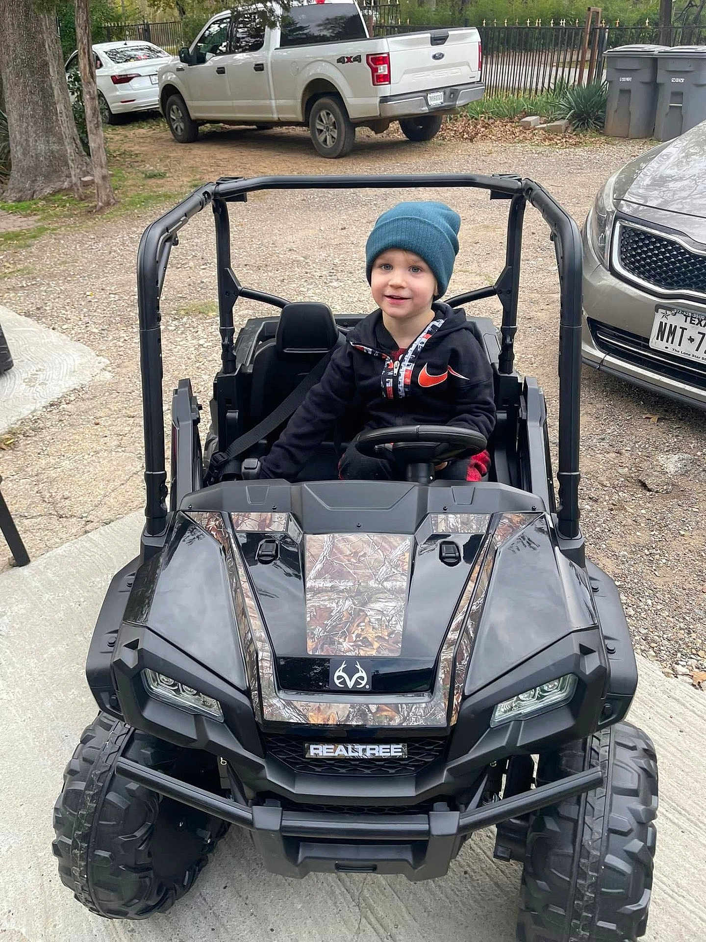 Zayden is registered to the contest to win money with this photo: child, toy_car, beanie, hoodie, outdoor, driveway, vehicle, smiling, parked, tree, car, wheel, tire, license_plate, gravel, concrete, person, casual_clothing, daylight, play