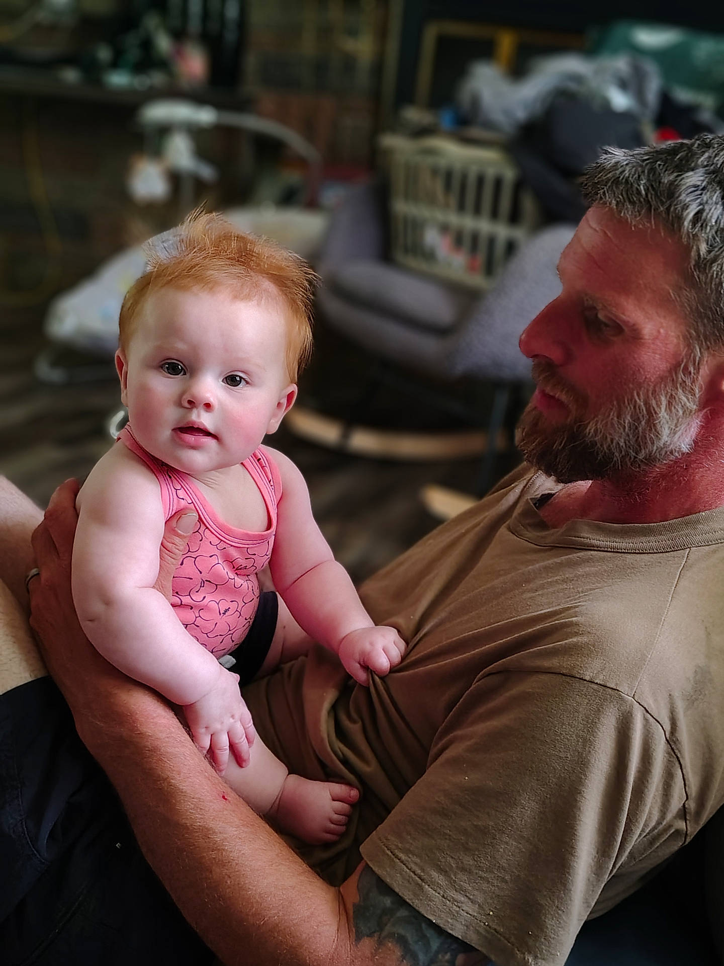 baby, blurred_background, bonding, casual_clothing, child, curly_hair, expression, facial_hair, family, holding, home, indoor, love, man, pink_tank_top, portrait, red_hair, seated, skin, tattoo