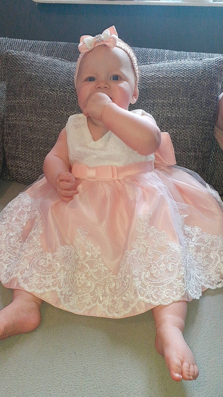 Mïa participe au concours pour gagner de l'argent avec cette photo : baby, baby_toddler_clothing, bridal_accessory, cheek, child, day_dress, dress, embellishment, happy, head, one_piece_garment, pattern, peach, person, pink, ruffle, sitting, skin, sleeve, toddler