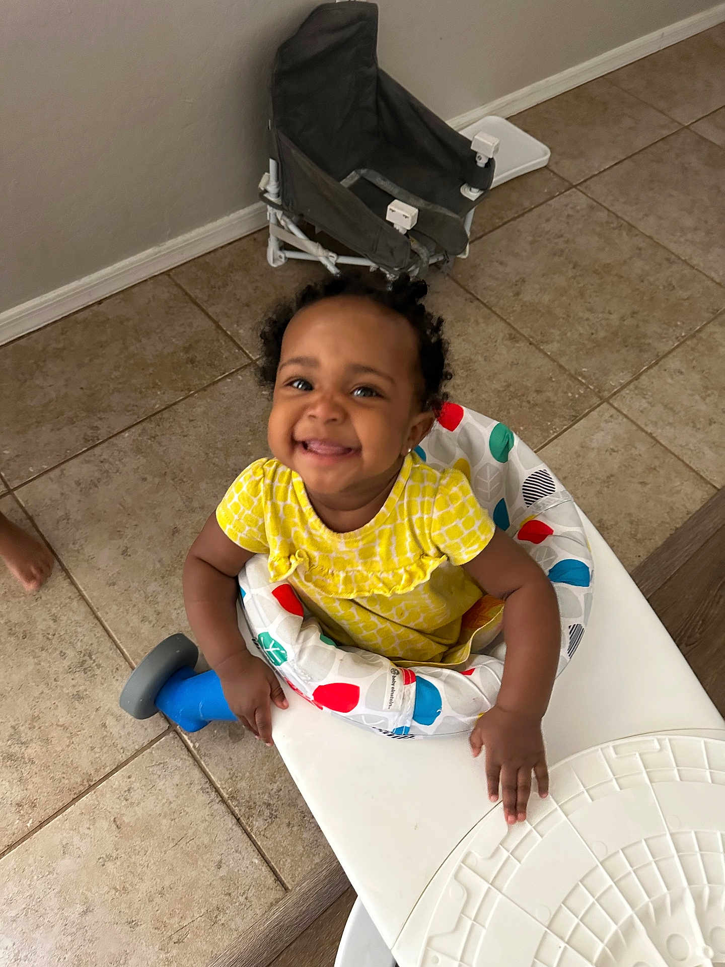Alaina is registered to the contest to win money with this photo: toddler, baby_walker, smiling, yellow_dress, indoor, tile_floor, child, happy, person, seat, playful, home, furniture, flooring, baby, cute, young_child, childhood, infant, clothing