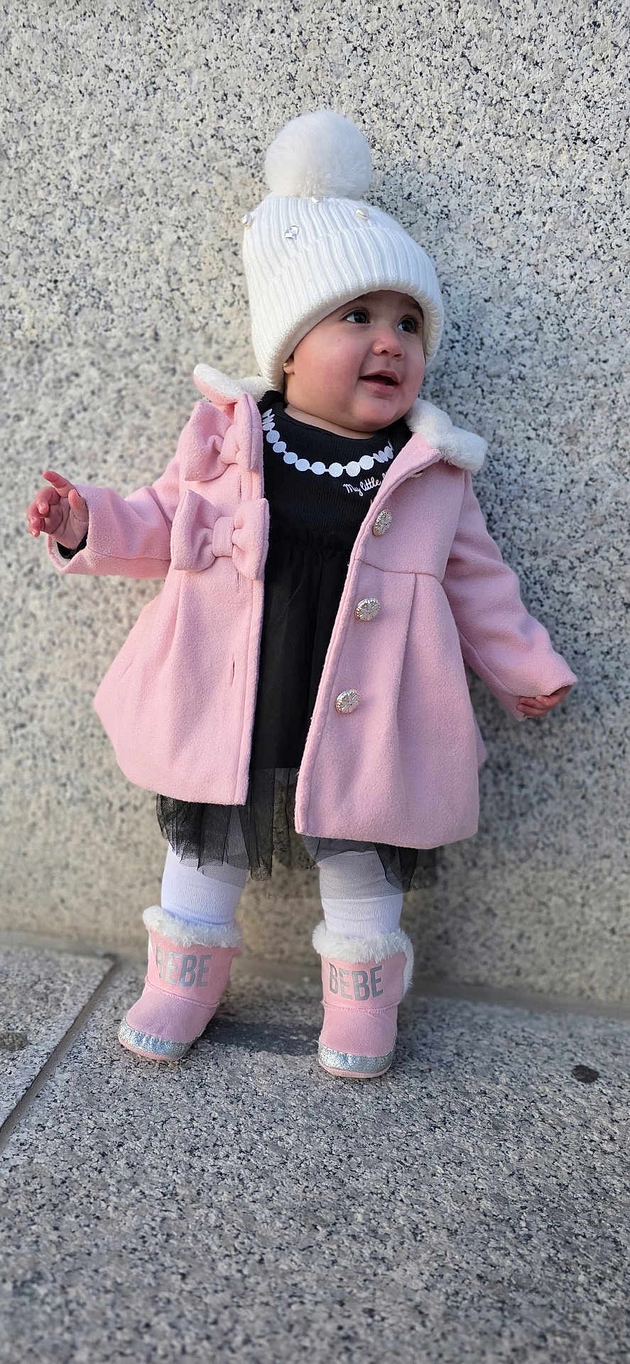 Aitana Sánchez joined the competition — help win amazing prizes! toddler, child, hat, pom_pom, pink_coat, bows, boots, outdoor, stone_wall, white_tights, black_dress, smiling, standing, winter_clothing, curious, portrait, footwear, baby, fashion, warm