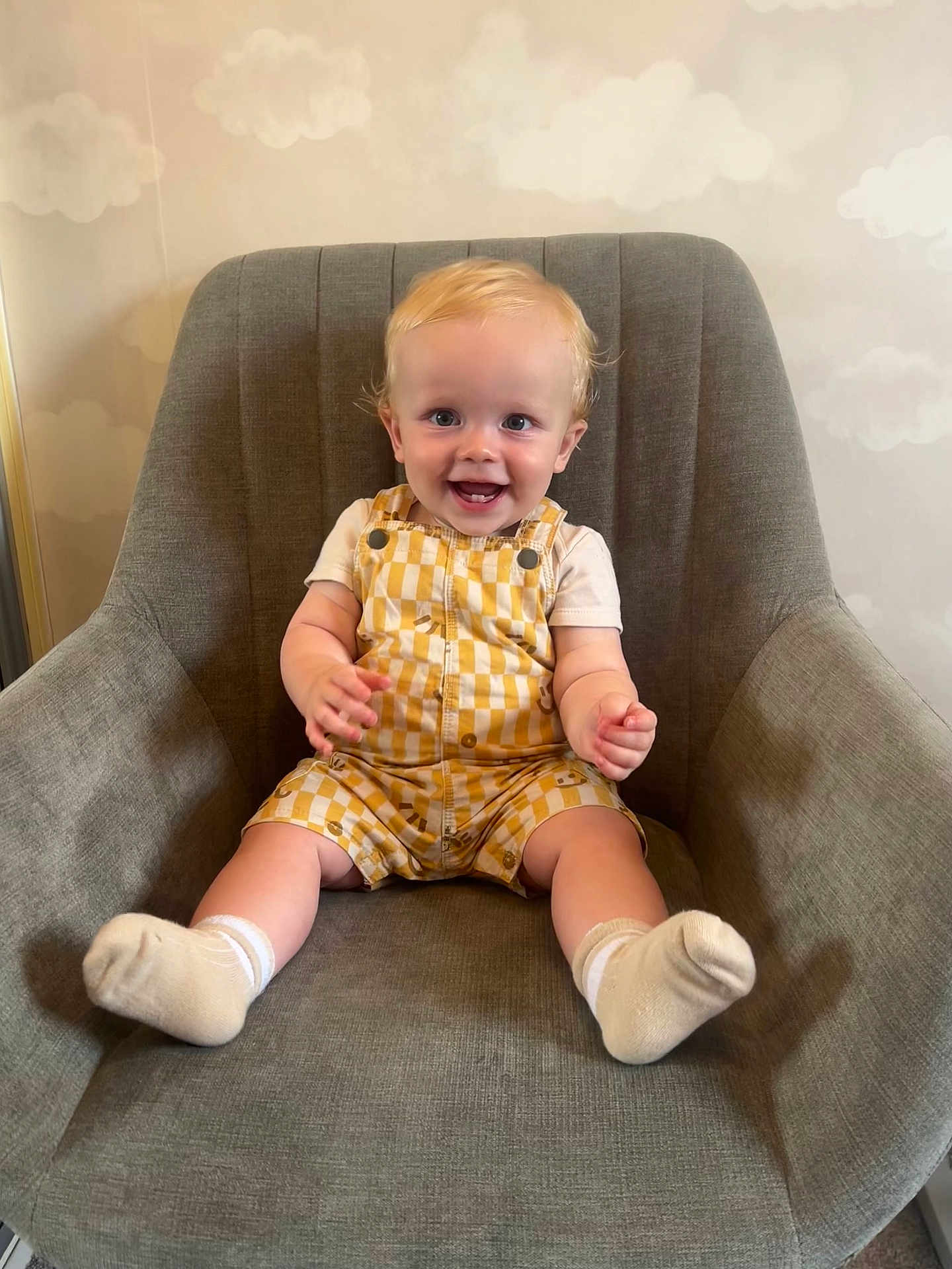 Bailey is registered to the contest to win money with this photo: toddler, child, smiling, yellow_overalls, white_socks, blond_hair, blue_eyes, grey_chair, indoor, portrait, happy, baby, cute, sitting, furniture, wallpaper, clouds, casual_clothing, human, home