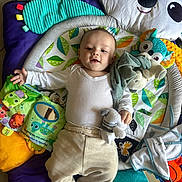 Jahlënn participe au concours pour gagner de l'argent avec cette photo : baby, child, infant, play_mat, soft_toys, stuffed_animal, colorful, smiling, clothing, white_shirt, beige_pants, mirror, floor_tiles, leaf_pattern, cute, happy, toy_book, plush, indoor, person