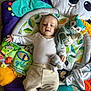 baby, infant, toy, stuffed_animal, play_mat, cushion, colorful, white_clothing, floor, happy, smiling, child, indoor, soft_toy, pillow, person, cute, comfort, leaves_pattern, relaxed