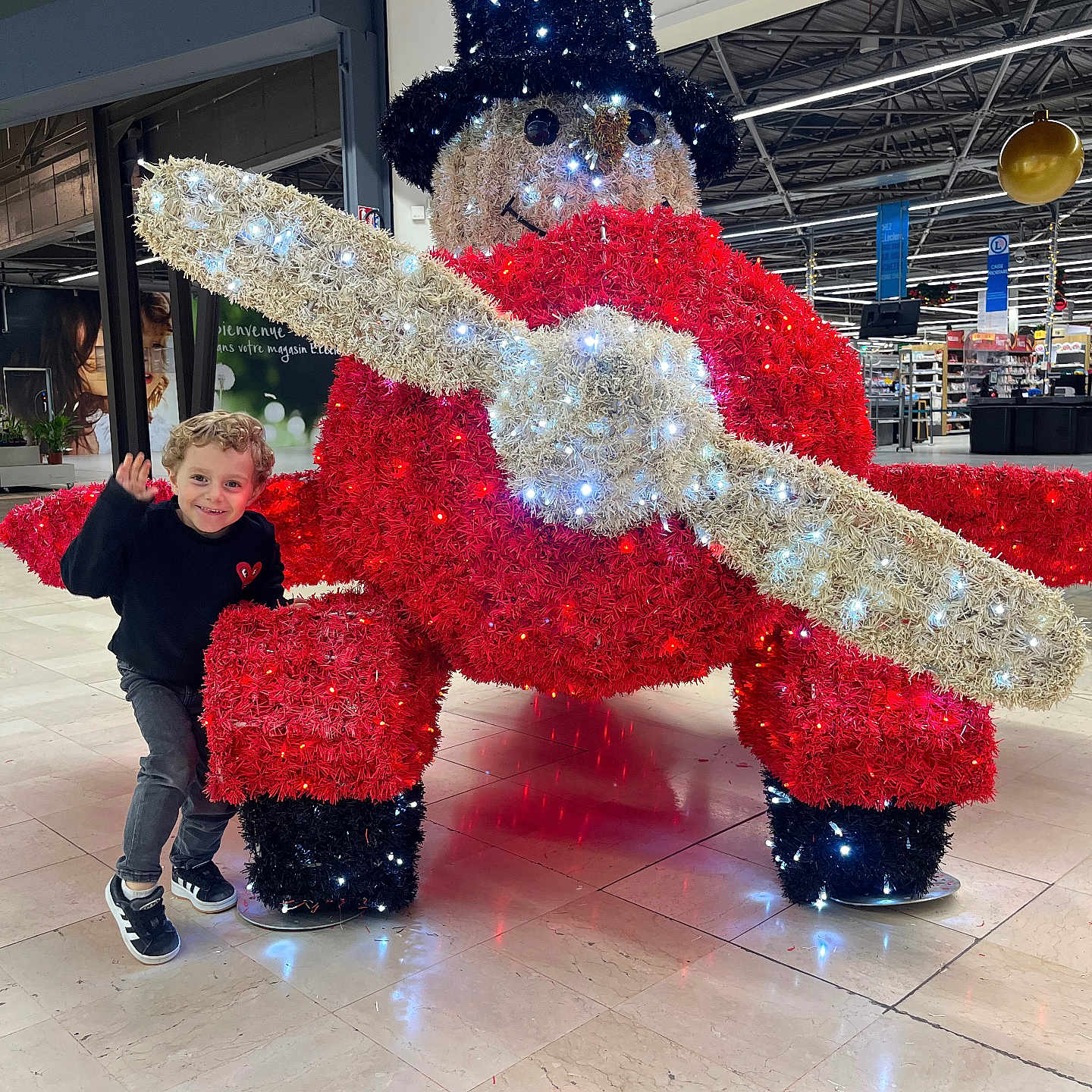 Petru participe au concours pour gagner de l'argent avec cette photo : child, smiling, waving, indoor, shopping_mall, festive_decoration, snowman, lights, top_hat, red, white, black, floor, tile_floor, propeller, holiday, decoration, person, happy, curly_hair