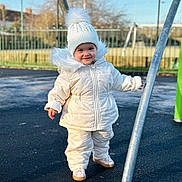 Rebecca joined the competition — help win amazing prizes! toddler, child, winter_clothing, white_jacket, white_hat, pom_pom, outdoor, playground, metal_pole, smiling, standing, park, fence, daylight, cold_weather, footwear, pavement, person, happy, cute