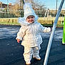toddler, child, winter_clothing, white_jacket, white_hat, pom_pom, outdoor, playground, metal_pole, smiling, standing, park, fence, daylight, cold_weather, footwear, pavement, person, happy, cute