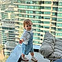toddler, child, balcony, denim_overalls, white_shirt, flower, short_hair, shoes, socks, modern_buildings, glass_railing, urban, outdoor, daylight, curious, standing, person, young_child, baby, casual_clothing
