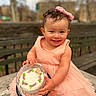 toddler, child, smile, dress, pink, bow, cake, plastic_cover, bench, outdoor, park, curly_hair, happy, sitting, cute, food, celebration, portrait, person, daylight