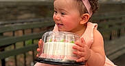 Grace is registered to the contest to win money with this photo: toddler, child, cake, pink_dress, headband, outdoor, bench, smile, happy, curly_hair, baby, holding, celebration, park, skin, person, cute, food_container, portrait, daylight