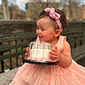 toddler, child, cake, pink_dress, headband, outdoor, bench, smile, happy, curly_hair, baby, holding, celebration, park, skin, person, cute, food_container, portrait, daylight