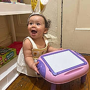 Grace is registered to the contest to win money with this photo: board_games, bunny_ears, child, curly_hair, dress, earrings, happy, home, indoor, monkey_around, person, pink, playtime, purple, shelf, smiling, toddler, toy_table, wooden_floor, yellow