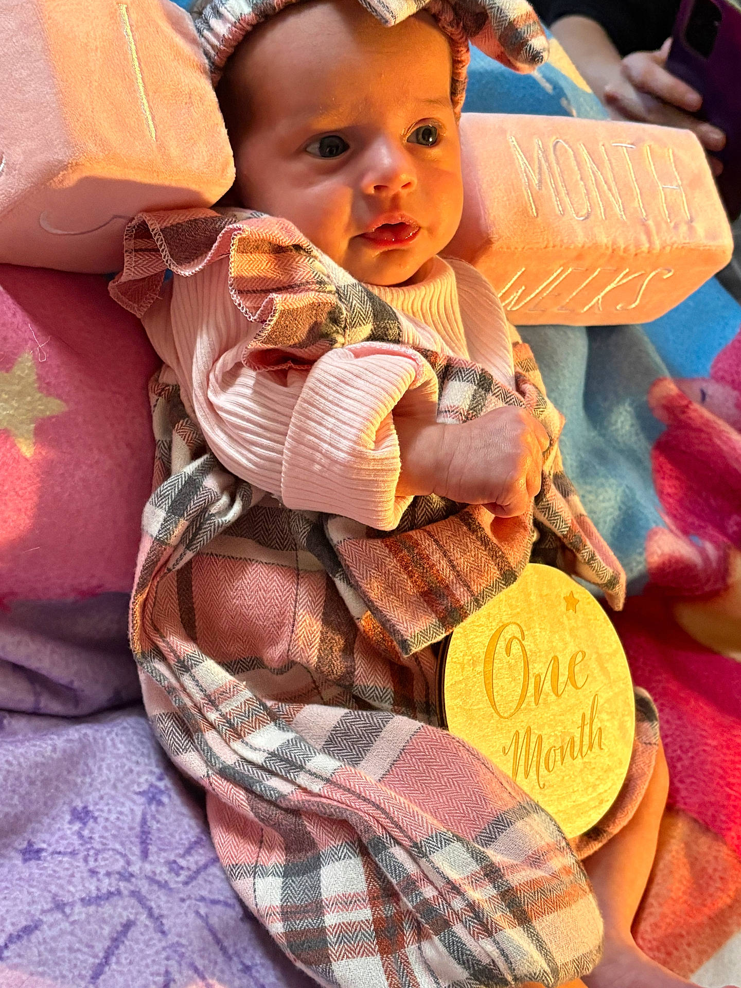 baby, child, colorful_blankets, cute, eyes, face, hand, indoors, infant, milestone, newborn, one_month, pink_block, plaid_clothing, portrait, sitting, soft_lighting, warm_colors, wooden_plaque, wrapped