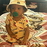 Royal joined the competition — help win amazing prizes! toddler, child, baby, pacifier, hat, bucket_hat, clothing, bed, blanket, indoor, person, face, sitting, pattern, books, furniture, curious, cute, young_child, cozy