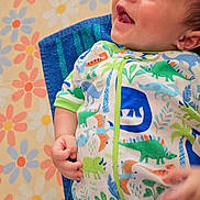 Emari is registered to the contest to win money with this photo: baby, infant, smiling, dinosaur_print, onesie, hand, face, happy, lying_down, blanket, floral_pattern, child, cute, skin, head, blue, green, orange, white, toddler