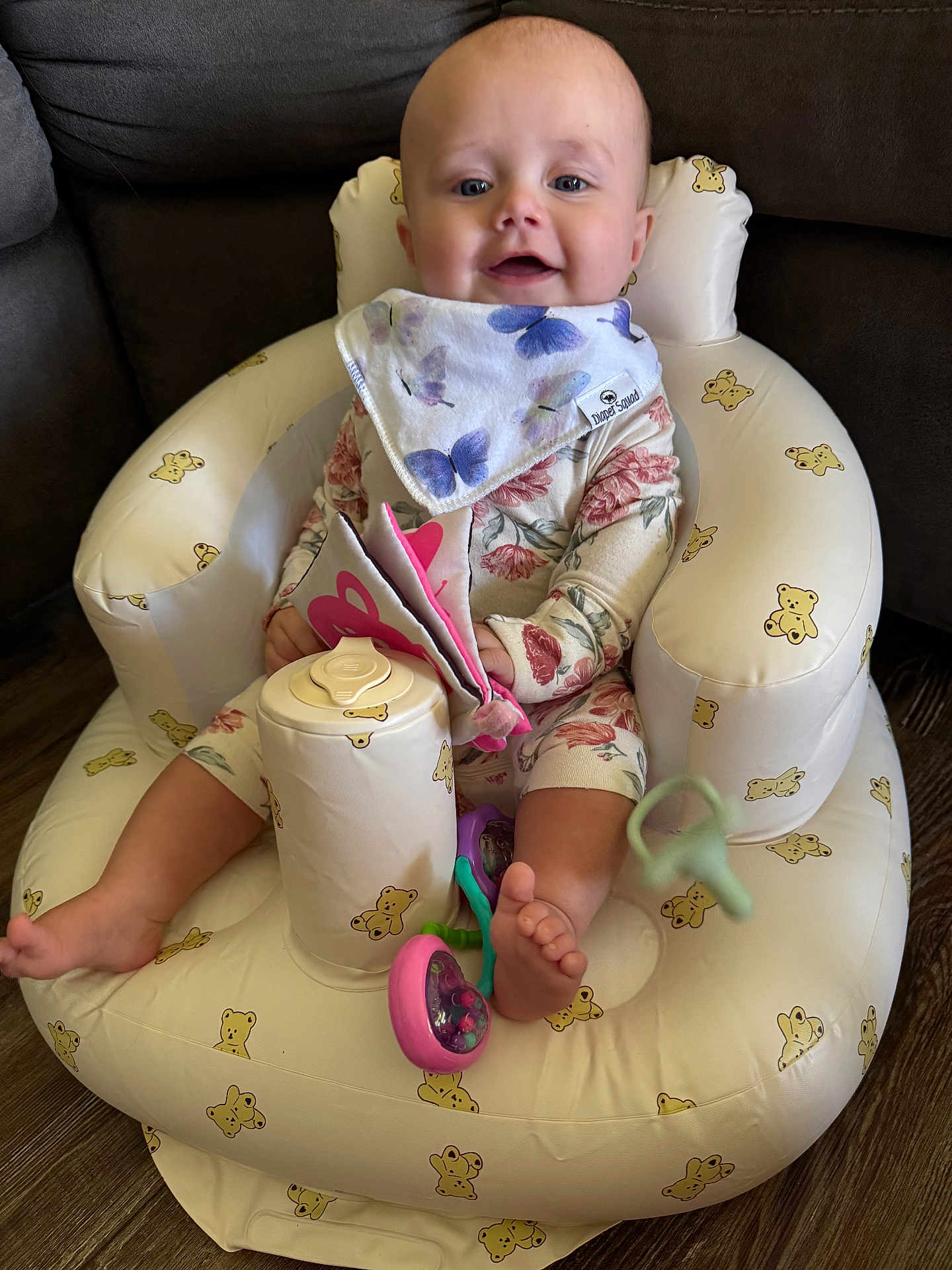 Patsy joined the competition — help win amazing prizes! baby, infant, child, inflatable_chair, teddy_bear_pattern, bib, floral_outfit, toy, soft_book, pink_toy, purple_toy, smiling, indoor, wood_floor, cute, seated, barefoot, happy, person, portrait