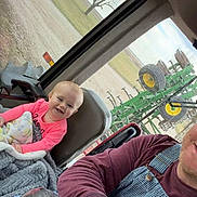 Patsy is registered to the contest to win money with this photo: baby, child, smile, infant, blanket, steering_wheel, tractor_cab, overalls, watch, adult, man, vehicle_interior, farm_equipment, field, tree, window, seat, denim, cozy, portrait