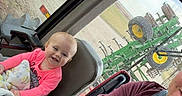 Patsy is registered to the contest to win money with this photo: adult, baby, blanket, child, cozy, denim, farm_equipment, field, infant, man, overalls, portrait, seat, smile, steering_wheel, tractor_cab, tree, vehicle_interior, watch, window