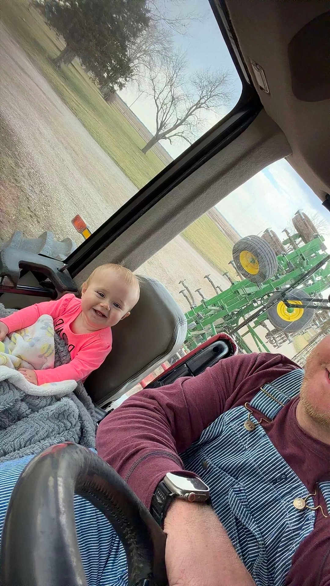 Patsy is registered to the contest to win money with this photo: baby, child, smile, infant, blanket, steering_wheel, tractor_cab, overalls, watch, adult, man, vehicle_interior, farm_equipment, field, tree, window, seat, denim, cozy, portrait