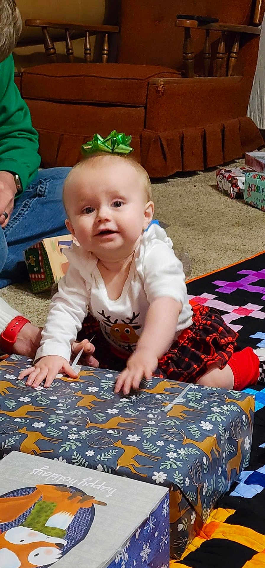 Patsy is registered to the contest to win money with this photo: baby, child, gift, present, wrapping_paper, holiday, christmas, reindeer, green_bow, carpet, quilt, plaid_skirt, sitting, indoor, person, cute, festive, holiday_decor, smiling, family