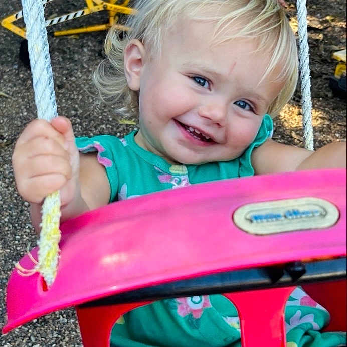 Patsy is registered to the contest to win money with this photo: blond_hair, blue_eyes, child, green_clothing, grinning, hands, happy, joy, outdoor, pebbles, pink_swing, play_equipment, playground, playtime, portrait, ropes, sitting, smiling, swing, toddler