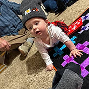 Patsy is registered to the contest to win money with this photo: baby, child, hat, knit_hat, crawling, quilt, carpet, floor, indoor, people, adult, sitting, plaid_skirt, long_sleeve, casual_clothing, family, curious, toy, patch, colorful