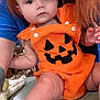 baby, child, pumpkin_outfit, orange_clothing, headband, bow, face, person, indoor, tattoo, arm, skin, table, furniture, hair, blue_shirt, cute, holiday, festive, expression