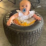 Patsy is registered to the contest to win money with this photo: baby, pacifier, tire, garage, concrete_floor, folding_chair, child, indoor, curious, clothing, playful, person, sitting, blue_eyes, white_shirt, yellow_skirt, toddler, floor, seat, object