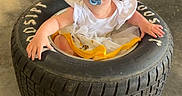 Patsy is registered to the contest to win money with this photo: baby, blue_eyes, child, clothing, concrete_floor, curious, floor, folding_chair, garage, indoor, object, pacifier, person, playful, seat, sitting, tire, toddler, white_shirt, yellow_skirt