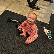 Patsy is registered to the contest to win money with this photo: baby, child, infant, pink_onesie, dumbbell, toy, rattle, floor_mat, gym_equipment, exercise, indoor, cute, playful, sitting, smiling, person, carpet, fitness, baby_toy, home