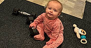Patsy is registered to the contest to win money with this photo: baby, baby_toy, carpet, child, cute, dumbbell, exercise, fitness, floor_mat, gym_equipment, home, indoor, infant, person, pink_onesie, playful, rattle, sitting, smiling, toy
