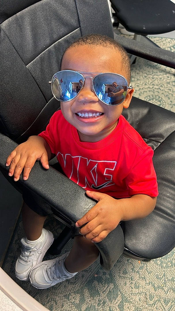 Zion is registered to the contest to win money with this photo: automotive_design, car_seat, comfort, cool, electric_blue, eyewear, finger, glasses, goggles, happy, joy, people, person, shoe, smile, sneakers, sunglasses, thumb, toddler, vehicle_door