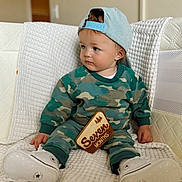 Callum is registered to the contest to win money with this photo: baby, child, infant, sitting, portrait, indoors, couch, blanket, quilt, baseball_cap, camouflage, sweatshirt, sneakers, milestone_sign, seven_months, wooden_sign, blue_eyes, serious_expression, furniture, family_photo