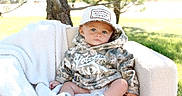 Callum is registered to the contest to win money with this photo: baby, child, infant, portrait, outdoor, sitting, blanket, chair, camouflage, hat, cap, clothing, shoes, white_shoes, grass, tree, nature, sunlight, serious_expression, close_up