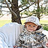 Callum is registered to the contest to win money with this photo: baby, child, infant, portrait, smiling, outdoor, pine_tree, nature, blanket, camouflage, hoodie, hat, clothing, grass, sunlight, backyard, sitting, lying_down, face, happy