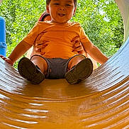 Leonardo is registered to the contest to win money with this photo: toddler, child, playground, slide, yellow_slide, orange_shirt, smiling, outdoor, trees, greenery, hands, adult, fun, play, summer, happy, person, park, casual_clothing, daytime
