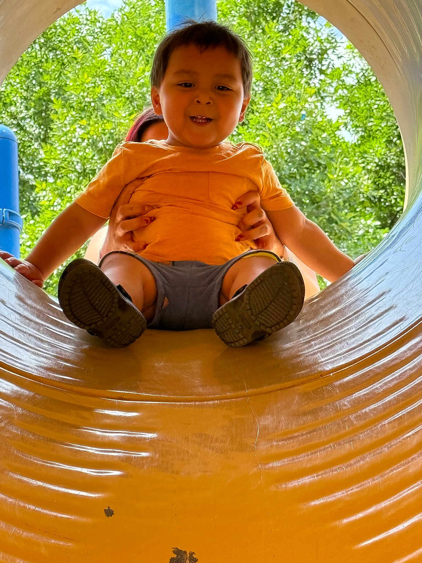 Leonardo is registered to the contest to win money with this photo: toddler, child, playground, slide, yellow_slide, orange_shirt, smiling, outdoor, trees, greenery, hands, adult, fun, play, summer, happy, person, park, casual_clothing, daytime