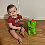 blue_eyes, camouflage_shorts, casual_clothing, child, curious, dinosaur, expression, floor, glowing_light, home, indoor, plastic_toy, playful, red_shirt, short_hair, sitting, toddler, toy, wooden_floor, young_child