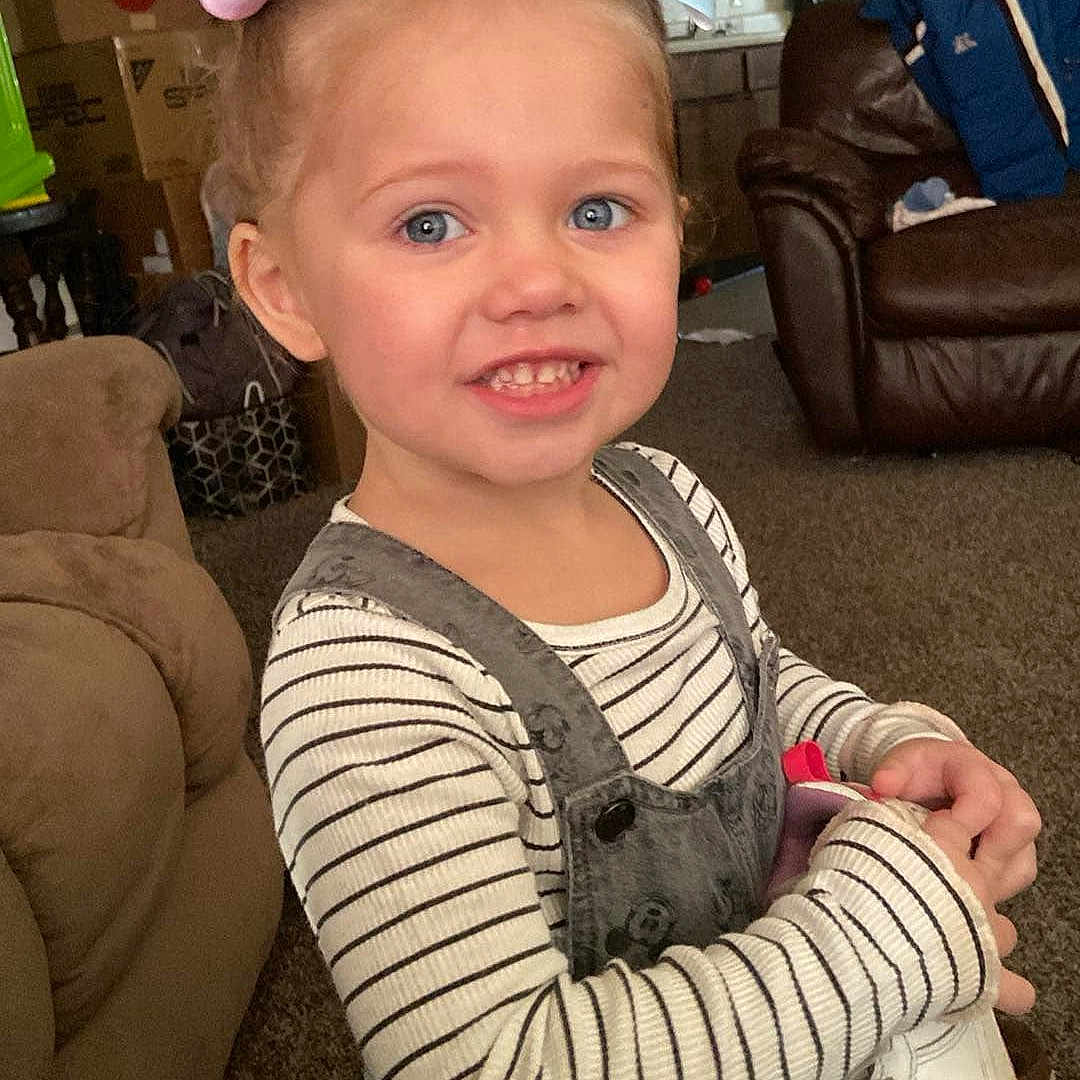 Delilah is registered to the contest to win money with this photo: blue_eyes, bow, boxes, carpet, casual_clothing, child, couch, furniture, girl, hair_accessory, happy, indoor, living_room, overalls, person, portrait, shoe, smiling, striped_shirt, toy