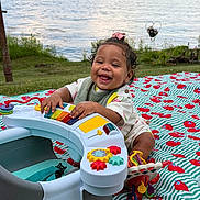 Hannah is registered to the contest to win money with this photo: baby, child, toy, piano, blanket, grass, lake, water, outdoor, smiling, happy, curly_hair, playtime, colorful, nature, greenery, summer, cute, infant, fun