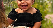 Hannah is registered to the contest to win money with this photo: adult, baby, black_dress, child, curly_hair, cute, daylight, earrings, greenery, happy, holding, joyful, nature, outdoor, person, plaid_shirt, portrait, red_bows, smiling, toddler