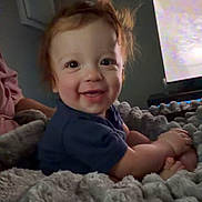 Jaden is registered to the contest to win money with this photo: baby, child, toddler, smiling, happy, portrait, sitting, indoor, blanket, fluffy_blanket, gray, blue_shirt, red_hair, face, eyes, television, living_room, cozy, home, photograph