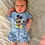 baby, infant, mickey_mouse, blue_clothing, romper, bedspread, patterned_bed, lying_down, cute, small_hands, small_feet, light_skin, curious_expression, indoors, soft_lighting, child, person, sleepwear, bedroom, resting