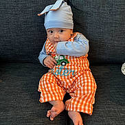 Benjamin is registered to the contest to win money with this photo: baby, child, infant, hat, blue_hat, orange_overalls, checkered_clothing, couch, sofa, furniture, sitting, indoors, cute, finger_in_mouth, wide_eyes, person, home, comfortable, plaid, casual_clothing