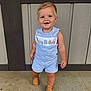 toddler, child, standing, romper, happy_birthday, blonde_hair, blue_eyes, smiling, footwear, loafers, concrete_floor, wooden_wall, outdoor, casual_clothing, cute, portrait, young_child, fashion, one_year_old, celebration