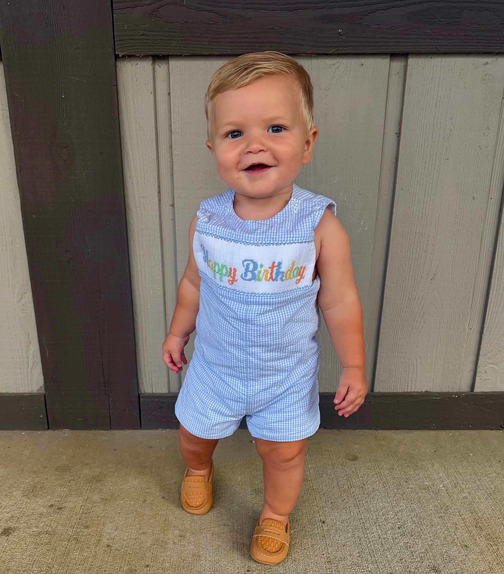 Beckett is registered to the contest to win money with this photo: toddler, child, standing, romper, happy_birthday, blonde_hair, blue_eyes, smiling, footwear, loafers, concrete_floor, wooden_wall, outdoor, casual_clothing, cute, portrait, young_child, fashion, one_year_old, celebration